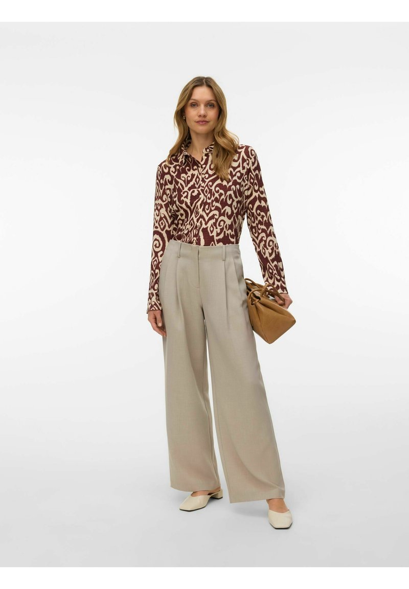 Femme portant une chemise à manches longues à motifs, un pantalon beige à jambes larges, des ballerines crème, et tenant un sac à main marron sur un fond uni.