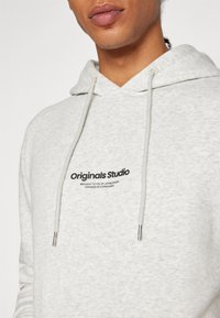 Felpa grigia chiara realizzata in tessuto morbido, con tasca frontale, caratterizzata da un logo nero "Originals Studio" e lacci con terminali metallici.