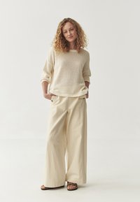 Pull en maille épaisse couleur crème avec des manches larges associé à un pantalon beige ample. Des sandales simples complètent le look. Design minimaliste.