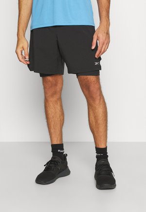 Mężczyzna w czarnych sportowych szortach z logo Reebok, czarnych skarpetkach Reebok i czarnych butach do biegania stojący na szarej podłodze.