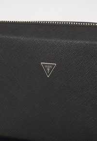 Cartera negra texturizada con un zipper plateado, que presenta un logo triádico de Guess USA en plata y negro en la parte frontal. Diseño simple y elegante.