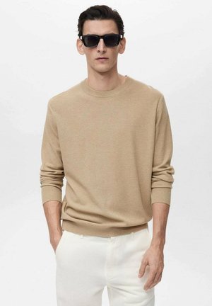 Beige knit sweater with round neckline and ribbed cuffs, paired with white pants. Man wearing black sunglasses stands against a light background.