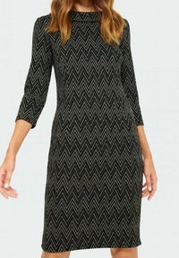 Žena v přiléhavých černých šatech s béžovým cik-cak vzorem a tříčtvrtečními rukávy, stojící s uvolněnýma rukama podél těla.