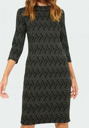 Femme portant une robe noire ajustée avec un motif zigzag beige et des manches trois-quarts, se tenant avec les bras relâchés sur les côtés.