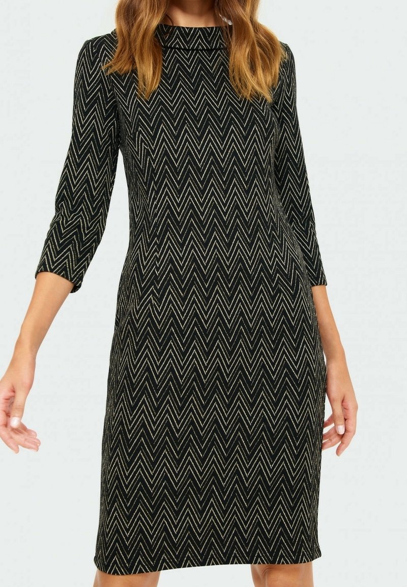 Žena v přiléhavých černých šatech s béžovým cik-cak vzorem a tříčtvrtečními rukávy, stojící s uvolněnýma rukama podél těla.