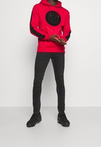 En person klädd i en röd hoodie med svarta ränder och emblem, svarta slim fit-jeans och svarta sneakers, som står framför en vit bakgrund.