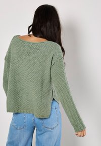 Grüner Strickpullover mit strukturiertem Muster, weitem Ausschnitt und langen Ärmeln. Kombiniert mit hellblauen Jeans, die eine lässige Passform zeigen.