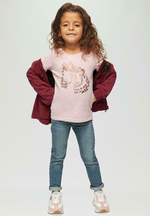 Helles rosa T-Shirt mit grafischem Design und braunem Text, weinroter Zip-Jacke, blaue Jeans und hellrosa Sneakers mit weißen Akzenten.