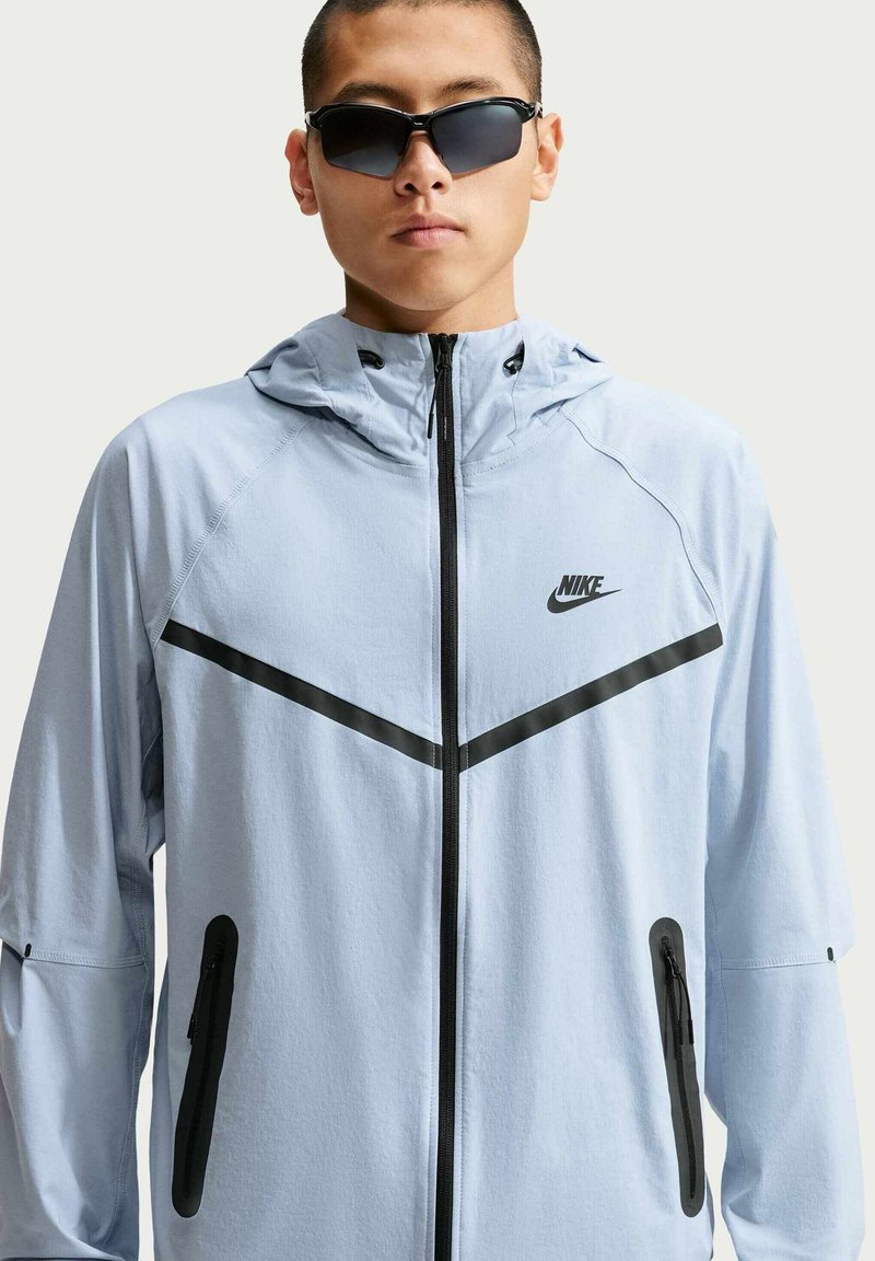 Mann trägt einen hellblauen Nike Windbreaker mit schwarzen Chevron-Streifen und Reißverschlusstaschen, trägt schwarze Umrandsonnenbrille.