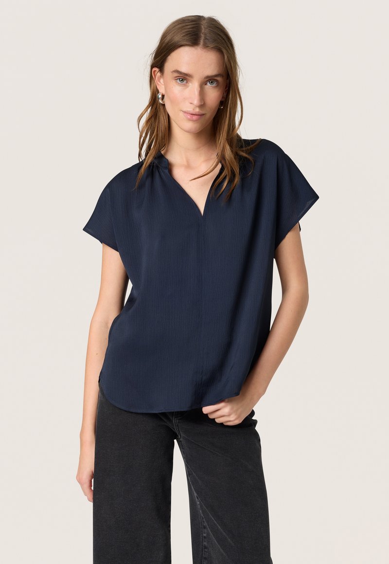 Blouse à manches courtes bleu marine avec un décolleté en V, fabriquée à partir de matériau texturé. Coupe large, fentes latérales et finition lisse. Associée à un pantalon noir.