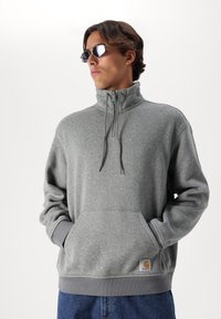 Sudadera gris claro de cuello alto, con cremallera en la parte delantera y bolsillo tipo canguro. Puños y dobladillo acanalados, parche de logo en el bolsillo.