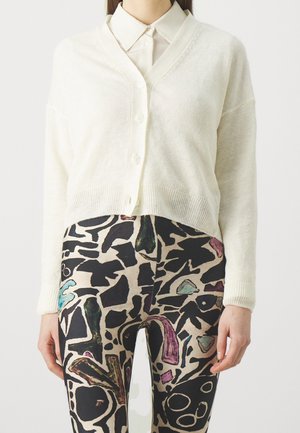 Personne portant un cardigan blanc boutonné sur une chemise beige avec des leggings à motifs présentant des formes abstraites noires et colorées.