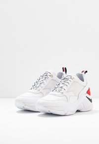 Baskets de sport blanches avec une tige en mesh et synthétique, bout rond, accents texturés, éclats noirs et rouges sur le talon et sur le côté.