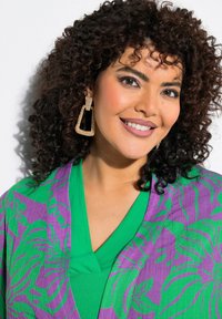 Bunter Kimono mit Blumenmuster in Lila und Grün, kombiniert mit einem einfarbigen grünen Oberteil; trägt große glitzernde geometrische Ohrringe.
