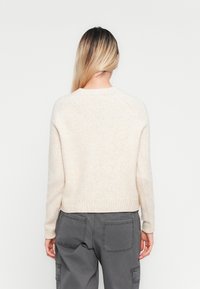 Una donna con lunghi capelli biondi indossa un maglione a maglia color crema e pantaloni cargo grigi, in piedi con la schiena rivolta verso la macchina fotografica su uno sfondo bianco.
