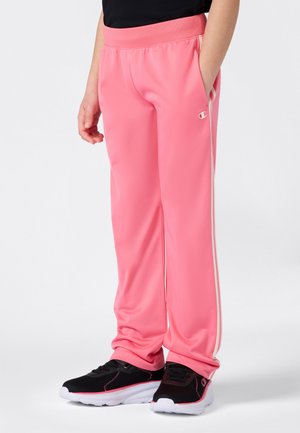 Une personne portant un pantalon de sport rose vif avec des rayures blanches sur les côtés et des baskets noires avec des semelles blanches, se tenant devant un fond uni.
