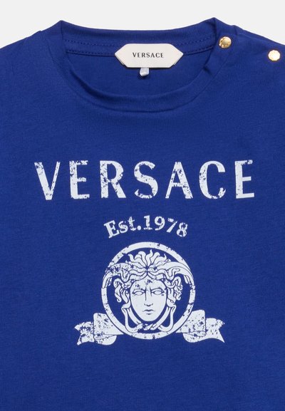 Μπλε πουκάμισο Versace με λευκό φθαρμένο λογότυπο, έμβλημα κεφαλής Μέδουσας και "Est. 1978" κάτω από το ντεκολτέ με δύο χρυσά κουμπιά στον ώμο.