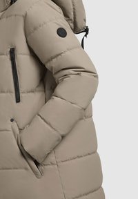 Beige pufferjakke med mat finish, der har vandret kviltning, en lynlåslomme på brystet og et sort logo-patch på ærmet.