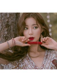 Un tubetto di rossetto rosso tenuto tra le mani, la modella indossa un vestito a fantasia floreale, bracciali sovrapposti e orecchini con frange, con onde morbide nei capelli.