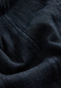 Mørkeblå denimstof med en let tekstureret overflade. Materialet fremstår blødt og krøllet, med en diskret glans og vævet mønster.