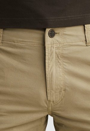 Close-up of beige pants with a visible button and stitching, worn with a dark brown shirt tucked in.