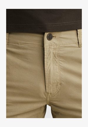 Close-up of beige pants with a visible button and stitching, worn with a dark brown shirt tucked in.