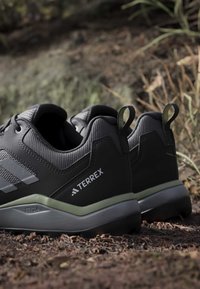 Chaussures de randonnée Adidas Terrex noires et grises reposant sur la terre d’un sentier forestier avec des feuilles sèches et un arrière-plan flou naturel.