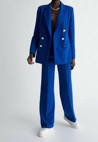 Niebieska dwurzędowa marynarka z złotymi guzikami, zestawiona z pasującymi szerokimi spodniami. Materiał wydaje się mieć teksturę; buty to białe trampki.
