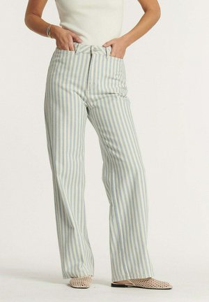 Kobieta ubrana w spodnie z wysokim stanem i prostymi nogawkami w pionowe niebiesko-białe paski, białą bezrękawnik i plecione płaskie buty.