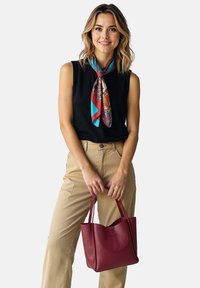 Crimson leather handbag with two handles, smooth texture, worn with beige pants and a black sleeveless top, accented by a colorful scarf.