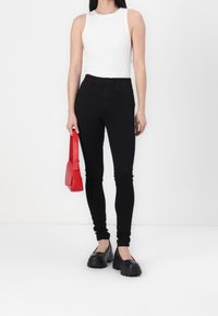 Black skinny jeans with a smooth texture, paired with a white ribbed tank top and chunky black slip-on shoes. Holding a small red handbag.