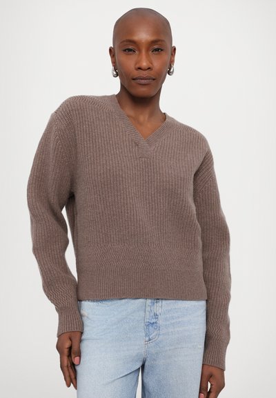 Strickjacke in taupe mit V-Ausschnitt, gerippter Textur und langen Ärmeln. Über hellblauen Jeans getragen, die eine lässige Passform zeigt.
