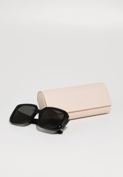 Occhiali da sole neri a forma quadrata con lenti scure, appoggiati accanto a un elegante astuccio rosa chiaro testurizzato con il nome del marchio "Chloé" stampato in rilievo.