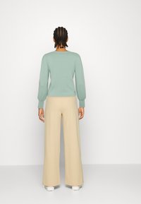 Hellgrüner Pullover mit langen, sanft gepufften Ärmeln und Rundhalsausschnitt, kombiniert mit beige-weiten Hosen und weißen Turnschuhen.
