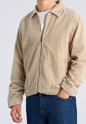 Mężczyzna w beżowej kurtce sztruksowej na suwak z elastycznym dołem i niebieskich dżinsach, z ręką w kieszeni na białym tle.