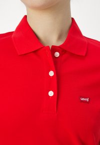 Camisa polo roja con cuello, tres botones blancos y un pequeño logo de Levi's en el pecho. Tejido texturizado.