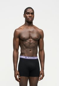 Μυώδης άνδρας στέκεται βλέποντας μπροστά, φορώντας μαύρα εσώρουχα μποξέρ με μωβ ζώνη μέσης που έχει την επιγραφή "BJÖRN BORG" σε απλό λευκό φόντο.