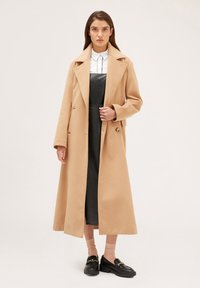 Beige dlouhý kabát s texturovaným povrchem, velký klopový límec, dva knoflíky, nošený přes černé šaty, v kombinaci s černými mokasíny a růžovými ponožkami.