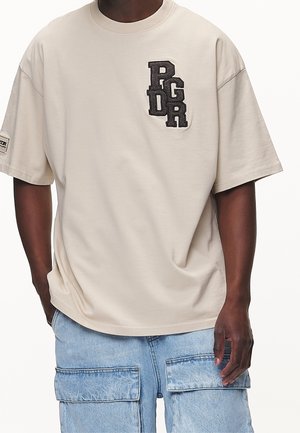 Homme portant un t-shirt beige oversize avec des lettres noires "PGRD" sur la poitrine et un pantalon cargo en denim bleu clair avec de grandes poches.
