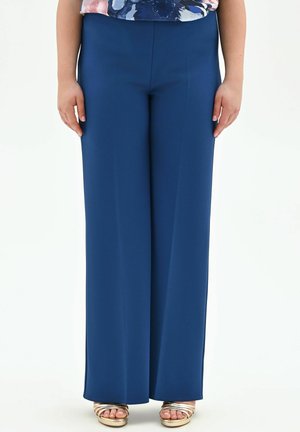 Femme portant un pantalon large taille haute bleu marine, des sandales ouvertes métalliques dorées, et un haut fleuri, debout devant un fond blanc.