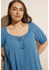 Blå, kortärmad blus med fyrkantig halsringning, samlad detaljer och en knytning fram. Lätt tyg med en slät textur.