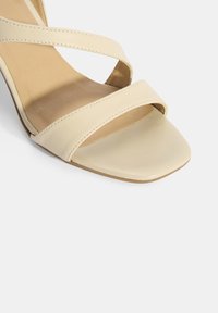 talon beige à lanières avec un orteil carré, fabriqué en cuir lisse, présentant une semelle intérieure douce et texturée et un design minimaliste.