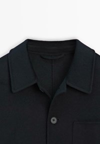 Mørkeblå skjortejakke i uldblandinger med flad krave, enkelt knaplukning og brystlomme. Blødt, struktureret stof med et minimalistisk design.