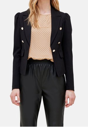 Woman wearing black double-breasted blazer with silver buttons, beige polka dot blouse, and black elastic-waist leather pants, no head shown.