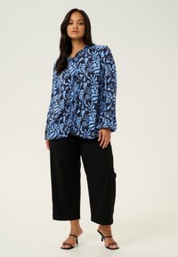 Bluse mit blauem Blumenmuster, rundem Ausschnitt und langen Ärmeln, kombiniert mit schwarzen weiten Hosen und schwarzen High-Heels.