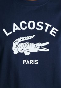 Tricou din bumbac bleumarin cu logo-ul alb al unui crocodil și textul "LACOSTE PARIS", cu guler rotund și mâneci scurte.