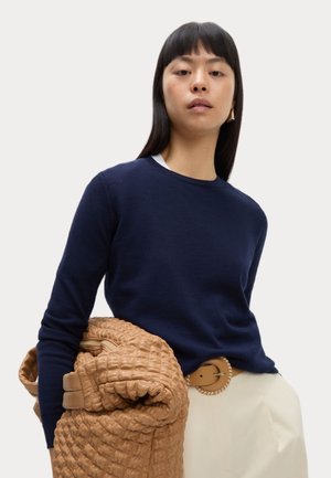 Mørkeblå strikket sweater med rund hals, iført en tan pufferjakke. Har et brunt bælte med en rund spænde og hvide bukser.