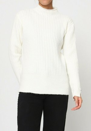 Pull blanc côtelé avec col montant, manches longues et coupe ajustée. Texture en maille, avec de subtils détails aux poignets. Porté avec un pantalon noir.