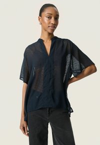Blusa blu navy a maniche corte con una texture leggera e trasparente e un colletto leggermente aperto. Presenta un delicato motivo a pois.