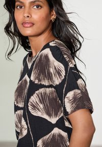 Black blouse featuring a large seashell print in beige, with short sleeves and a round neckline. The fabric appears lightweight and textured.
